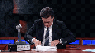 A Man In A Suit And Tie Sits At A Table With A Microphone GIF