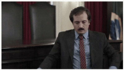 A Man In A Suit And Tie Sits In A Chair GIF
