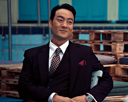 A Man In A Suit And Tie Sits In A Chair With His Hands Folded GIF