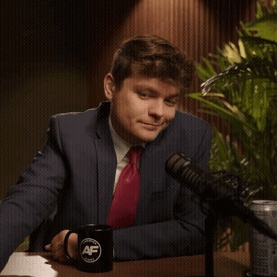 A Man In A Suit And Tie Sits In Front Of A Microphone With A Mug That Says Af GIF