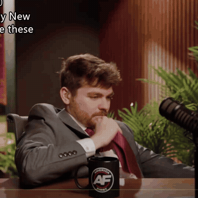 A Man In A Suit And Tie Sits In Front Of A Microphone With A Mug That Says " America First " On It GIF