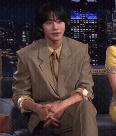 A Man In A Suit And Tie Sits On A Couch With His Hands Folded GIF