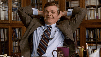 A Man In A Suit And Tie Sits With His Hands Behind His Head In Front Of A Bookshelf GIF