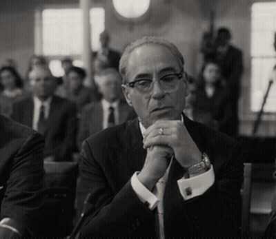 A Man In A Suit And Tie Sits With His Hands Folded In Front Of Him GIF