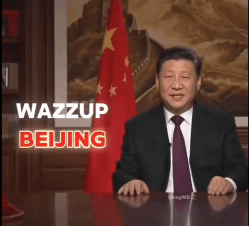 A Man In A Suit And Tie Sitting At A Desk With The Words Wazzup Beijing Behind Him GIF