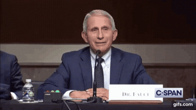 A Man In A Suit And Tie Sitting At A Table With A Sign That Says Dr. Fauci GIF