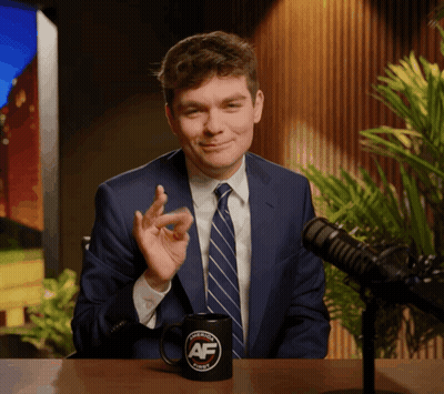 A Man In A Suit And Tie Sitting In Front Of A Microphone With A Mug That Says Avenue Af On It GIF