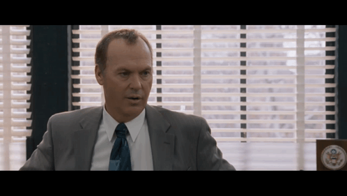 A Man In A Suit And Tie Sitting In Front Of A Window GIF