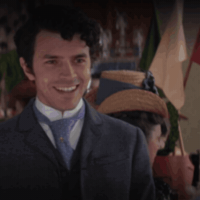 A Man In A Suit And Tie Smiles In Front Of A Woman In A Straw Hat GIF