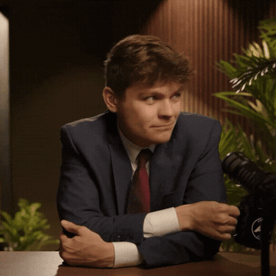 A Man In A Suit And Tie Smiles While Sitting At A Table With A Microphone In The Background GIF