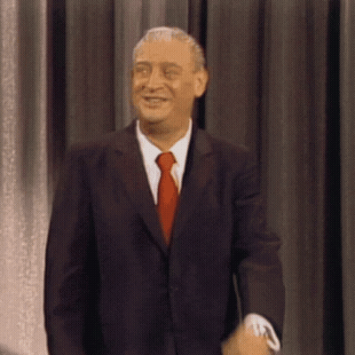 A Man In A Suit And Tie Smiles While Standing In Front Of A Curtain GIF