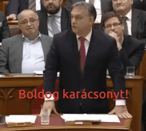 A Man In A Suit And Tie Stands At A Podium With The Words Boldog Karacsonyt In Red Behind Him GIF
