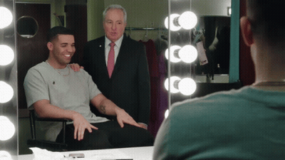 A Man In A Suit And Tie Stands Behind A Man In A Chair GIF