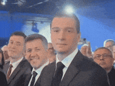A Man In A Suit And Tie Stands In A Crowd GIF