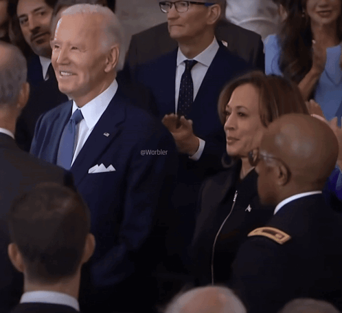 A Man In A Suit And Tie Stands In A Crowd With The Word Warbler On The Bottom Left GIF