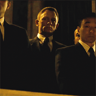 A Man In A Suit And Tie Stands In A Dark Room With Other Men GIF