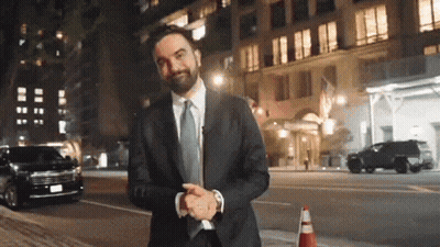 A Man In A Suit And Tie Stands In Front Of A Building At Night GIF