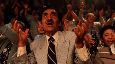 A Man In A Suit And Tie Stands In Front Of A Crowd With His Hands In The Air GIF