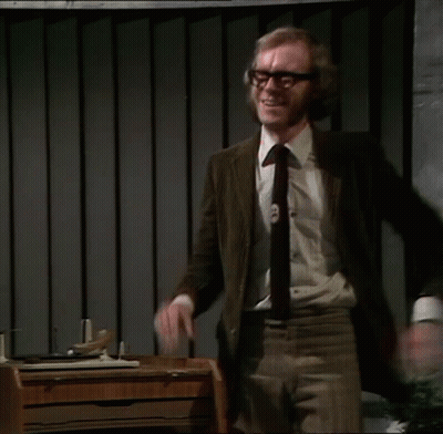 A Man In A Suit And Tie Stands In Front Of A Desk With His Arms Outstretched GIF