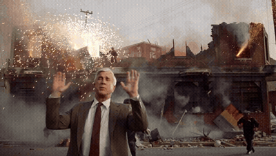 A Man In A Suit And Tie Stands In Front Of A Destroyed Building GIF