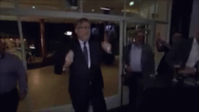 A Man In A Suit And Tie Stands In Front Of A Group Of People And Says " Voetjes Van De Vloer " In Yellow Letters GIF