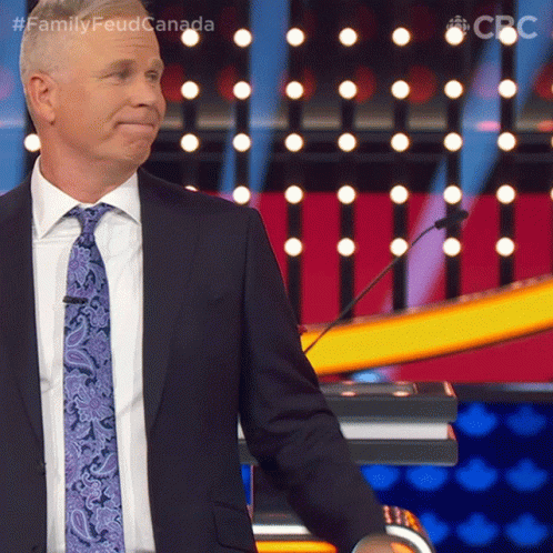 A Man In A Suit And Tie Stands In Front Of A Podium With The Words Family Feud Canada Written Above Him GIF