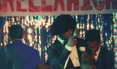 A Man In A Suit And Tie Stands In Front Of A Sign That Says " All-American " GIF