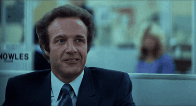 A Man In A Suit And Tie Stands In Front Of A Sign That Says Nowles GIF