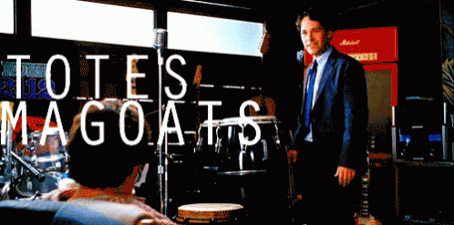 A Man In A Suit And Tie Stands In Front Of A Sign That Says Tote 'S Magoats GIF