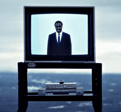 A Man In A Suit And Tie Stands In Front Of A Television That Says Lumin On It GIF