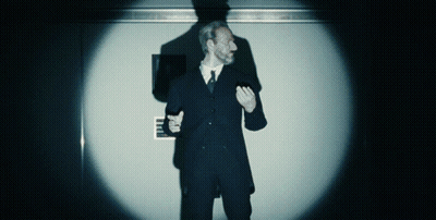 A Man In A Suit And Tie Stands In Front Of A Wall With A Sign That Says ' Emergency Exit ' On It GIF