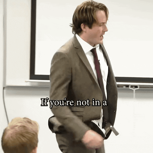 A Man In A Suit And Tie Stands In Front Of A White Board With The Words If You 'Re Not In A Written On It GIF