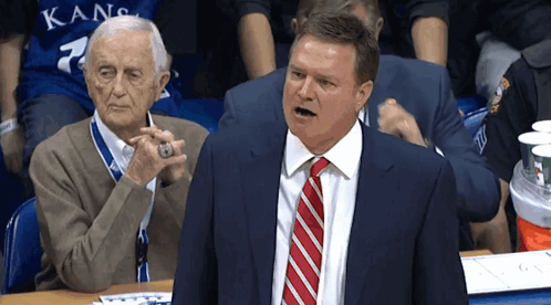 A Man In A Suit And Tie Stands In Front Of An Older Man In A Kansas Jersey GIF