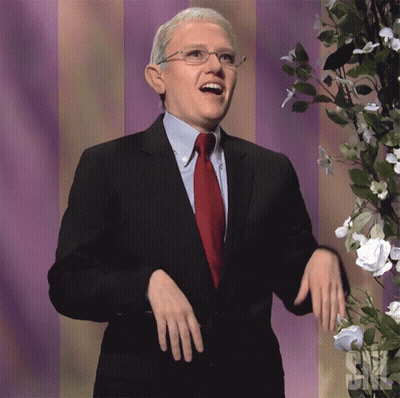 A Man In A Suit And Tie Stands In Front Of Flowers And A Sign That Says ' Snl ' GIF