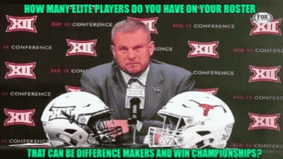 A Man In A Suit And Tie Stands In Front Of Two Football Helmets With Xii On Them GIF