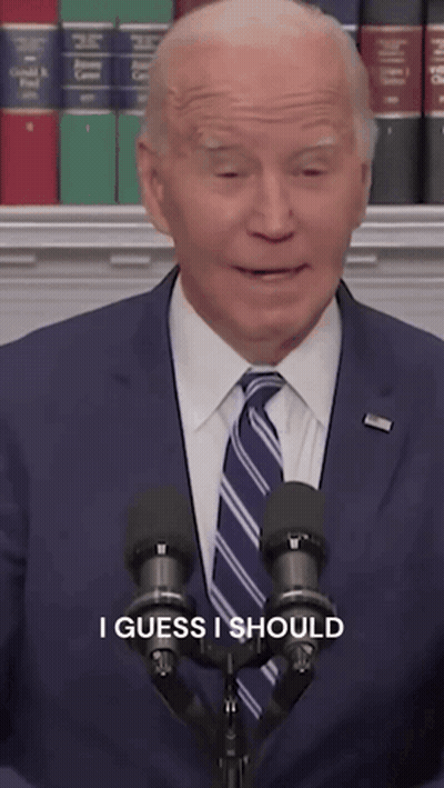 A Man In A Suit And Tie Stands In Front Of Two Microphones With The Words I Guess I Should Written Below Him GIF