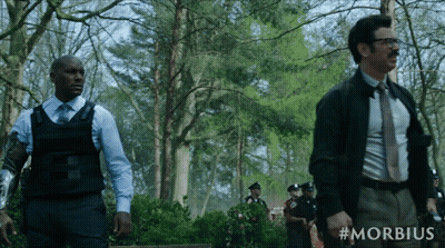 A Man In A Suit And Tie Stands Next To A Man In A Bullet Proof Vest In A Forest GIF
