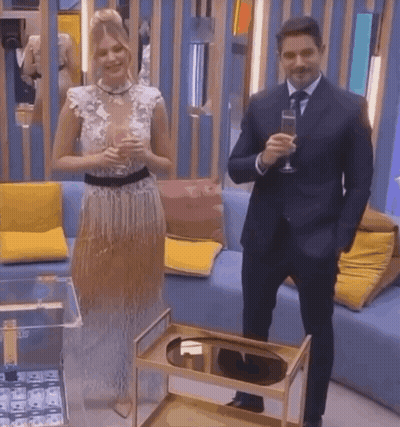 A Man In A Suit And Tie Stands Next To A Woman In A Dress Holding A Glass Of Champagne GIF