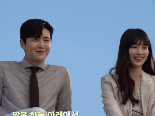 A Man In A Suit And Tie Stands Next To A Woman In A Polka Dot Jacket GIF