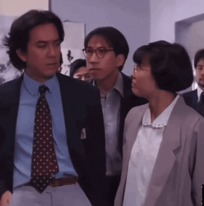 A Man In A Suit And Tie Stands Next To A Woman With Glasses GIF
