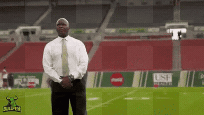 A Man In A Suit And Tie Stands On A Football Field With A Bullpen Logo On The Bottom GIF