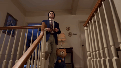 A Man In A Suit And Tie Stands On The Stairs Next To A Teddy Bear GIF
