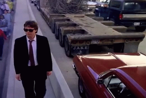 A Man In A Suit And Tie Walks Past A Truck That Says Jake On The Back GIF