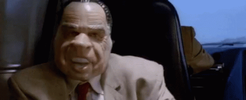 A Man In A Suit And Tie Wearing A Mask Is Sitting In A Chair . GIF
