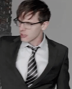 A Man In A Suit And Tie With Glasses Is Standing In A Room . GIF