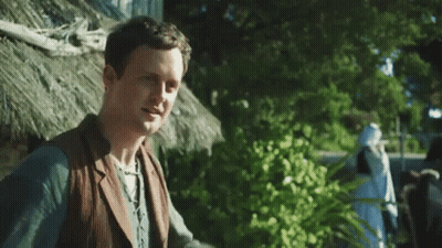 A Man In A Suit And Vest Is Standing In Front Of A Thatched Hut . GIF