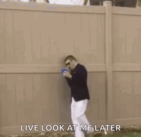 A Man In A Suit And White Pants Is Standing In The Grass Holding A Blue Cup . GIF