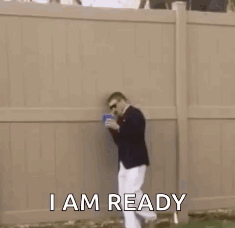 A Man In A Suit And White Pants Is Standing In The Grass With A Cup In His Hand . GIF