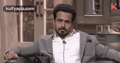 A Man In A Suit And White Shirt Is Sitting In A Chair With His Hands On His Knees . GIF