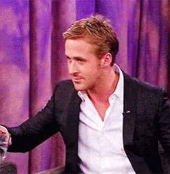 A Man In A Suit And White Shirt Is Sitting In Front Of A Purple Curtain And Holding A Glass Of Water . GIF